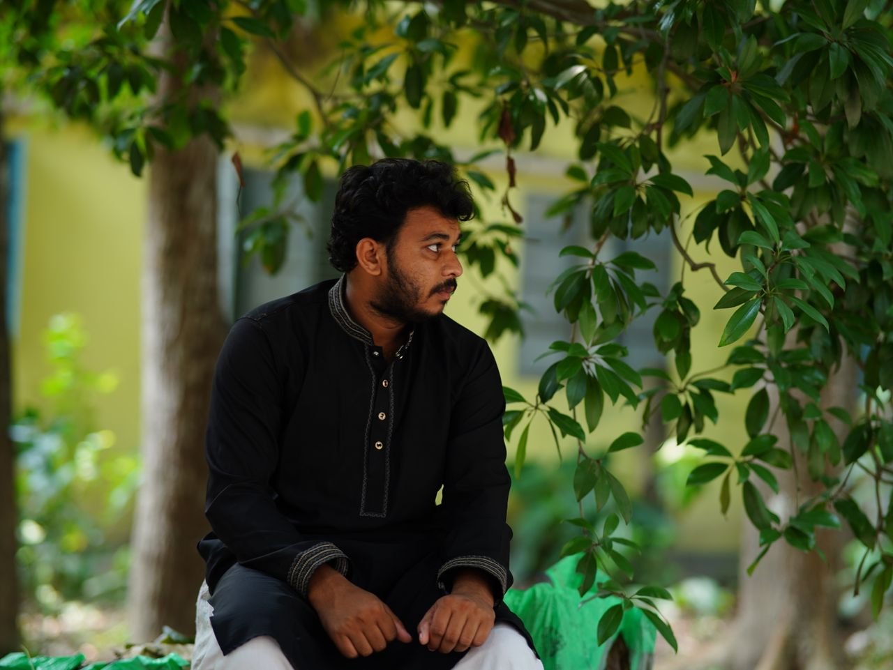Young man in traditional attire seated outdoors, shaded by lush greenery.