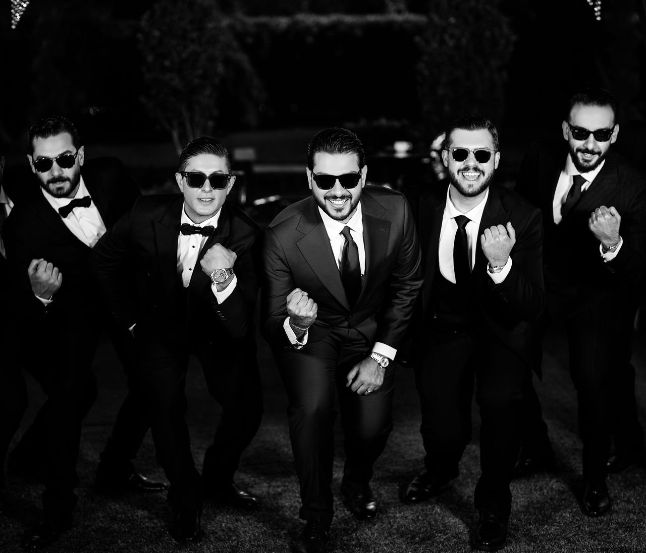 Group of groomsmen in suits and sunglasses posing energetically.