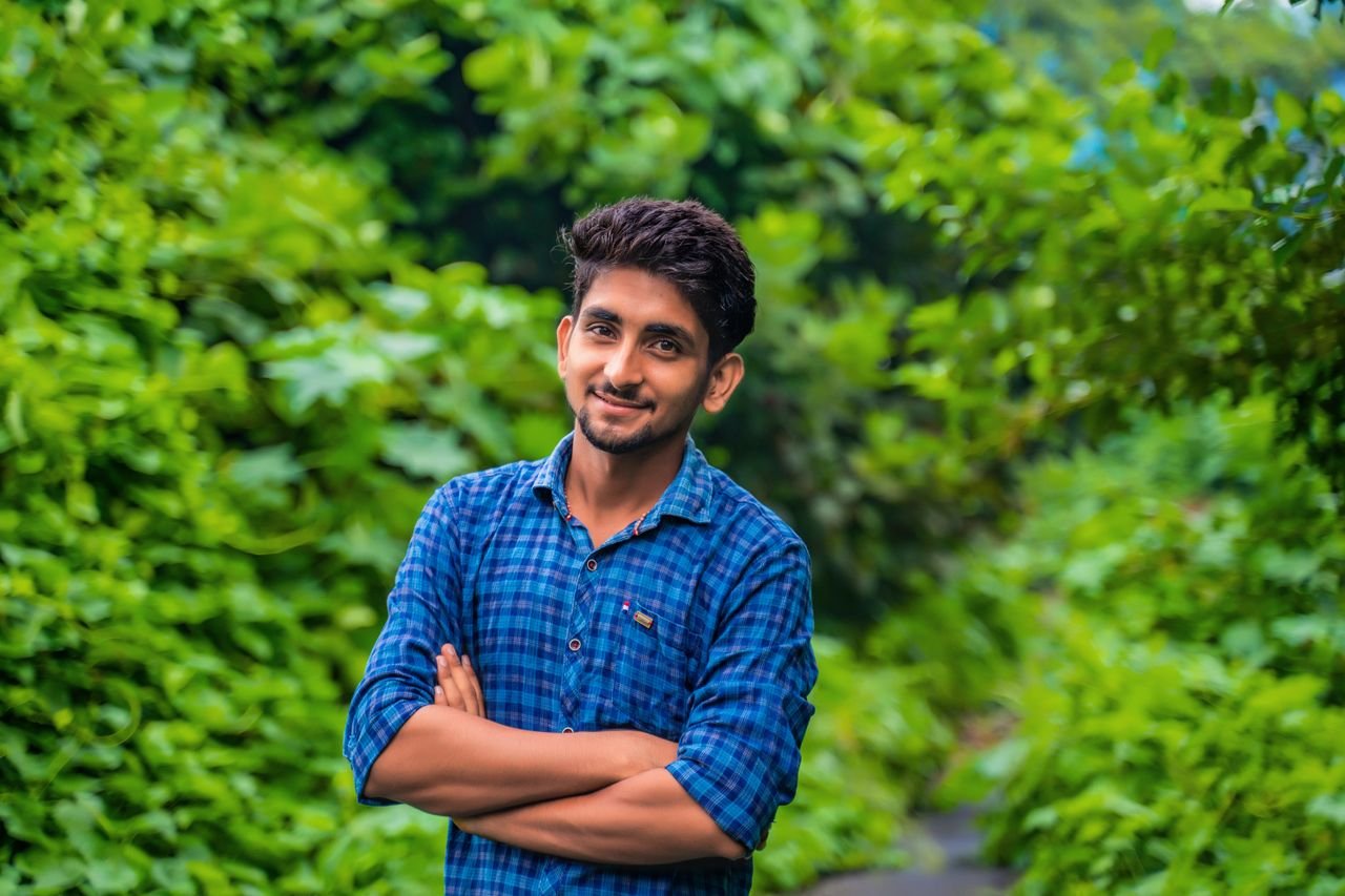 Young man in a checkered shirt with arms crossed, smiling against a lush green background.
